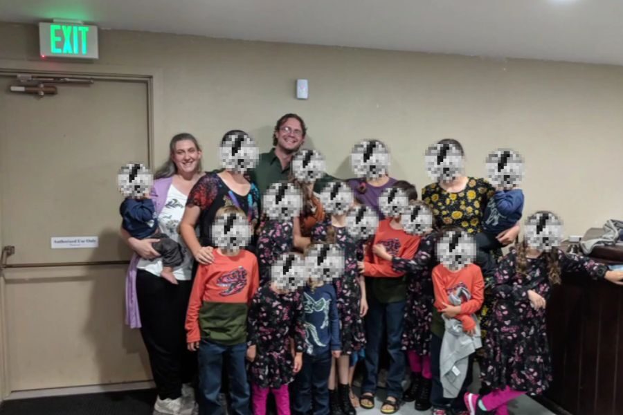 A large family poses together indoors near an exit door, with adults standing in the back and many children in front. The children's faces are pixelated for privacy.