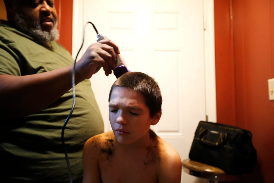 A man giving a young boy a haircut using clippers in a warmly lit room; the boy appears to be squinting.