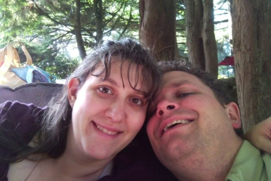 A couple smiles while leaning in close under the shade of tall trees during what appears to be a forest outing or picnic.