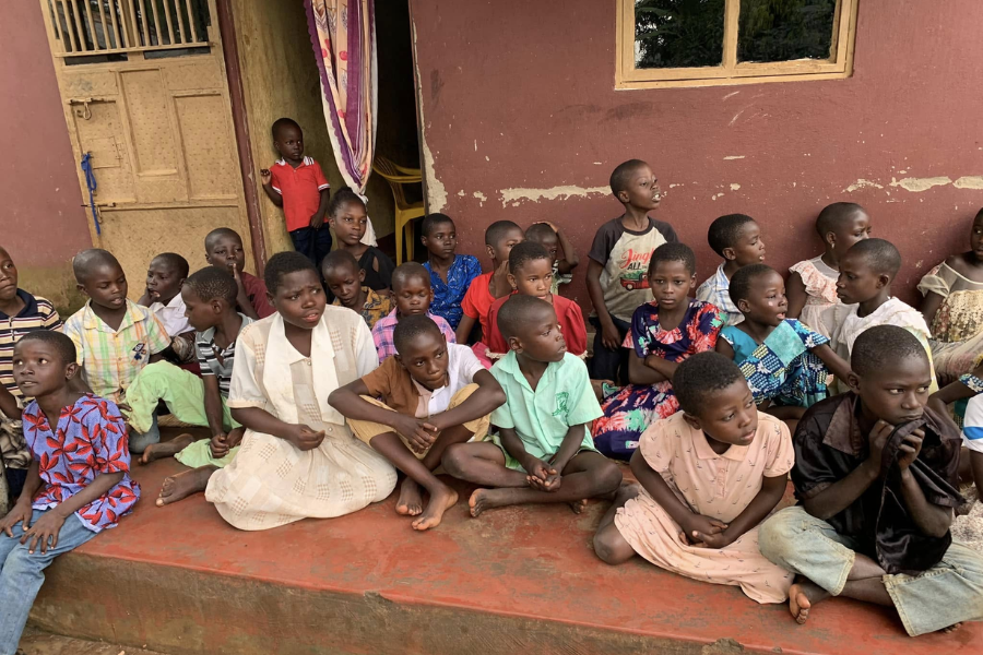 A group of children sitting on the porch of a worn building, some facing forward while others talk among themselves.
