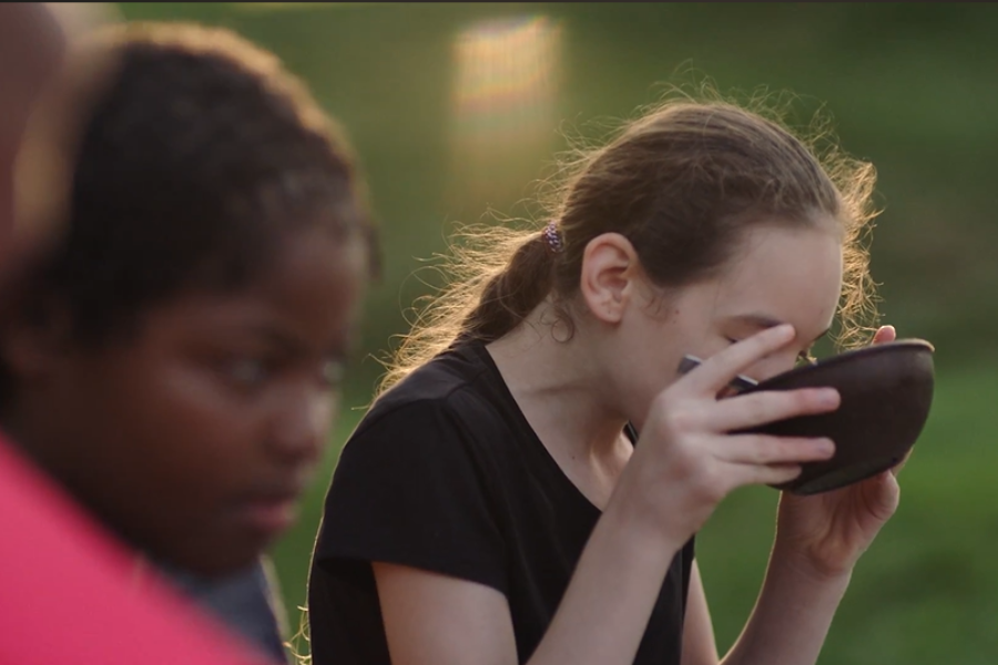 A girl drinking from a bowl outdoors while another child is seen slightly blurred in the foreground.