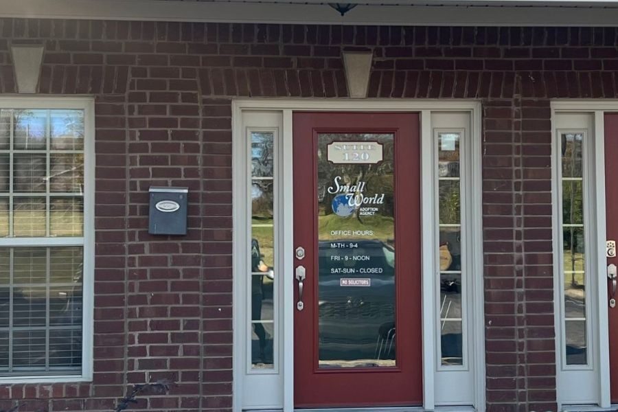 Glass door of “Small World Adoption Agency” office with business hours listed and “No Solicitors” sign, surrounded by red brick and two side windows.