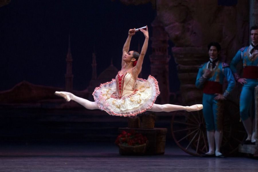 Ballerina in an ornate dress performs an elegant leap on stage, holding a fan, with two male dancers in colorful costumes watching in the background.