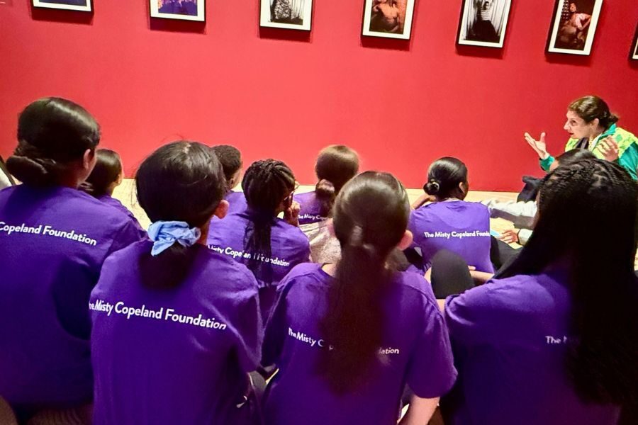 A group of children in purple "The Misty Copeland Foundation" shirts listens intently to a woman gesturing, seated against a red wall with framed photos.