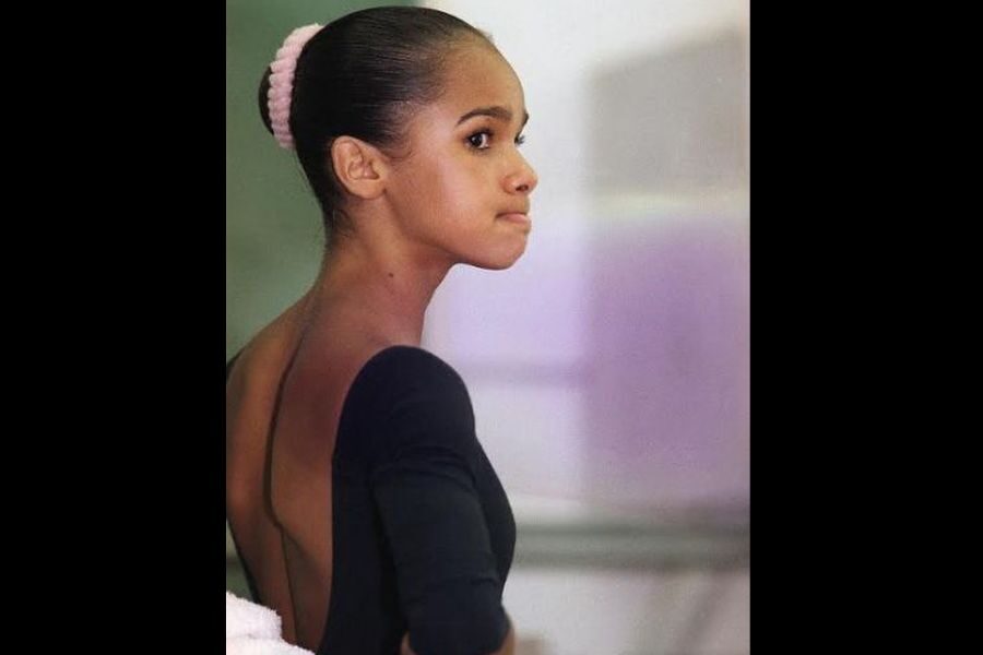 A young ballerina in a black leotard gazes thoughtfully off to the side. Her hair is slicked back in a bun with a pink hair accessory, conveying focus.