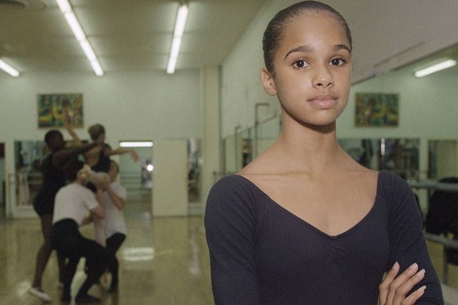 Young ballerina in a black leotard stands confidently with arms crossed in a dance studio. Dancers practice in the blurred background.