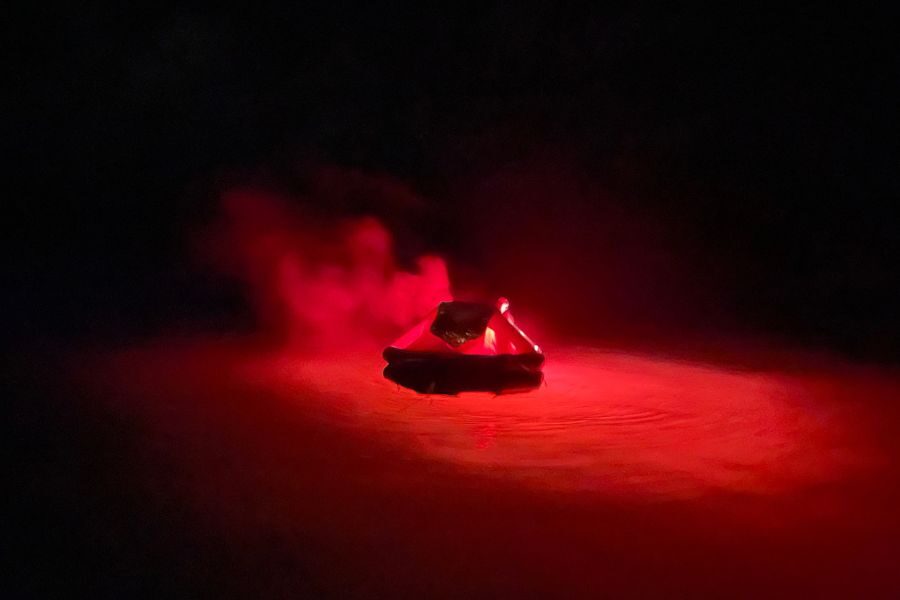 A person rides an inflatable tube in darkness, illuminated by a vivid red light that creates an eerie atmosphere. Red smoke adds a dramatic effect.