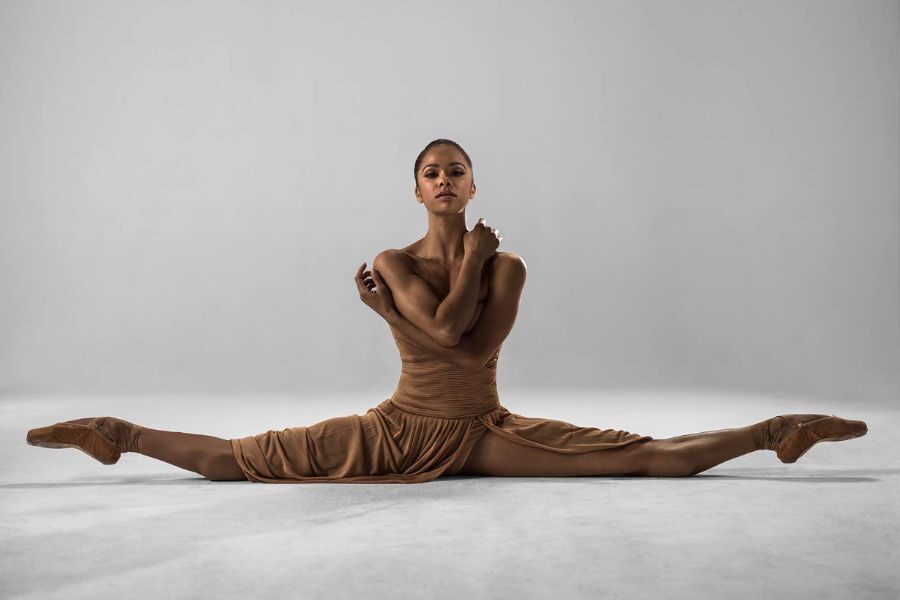 Ballet dancer in a tan dress performs a split on a gray background, arms gracefully crossed over her chest, conveying elegance and strength.
