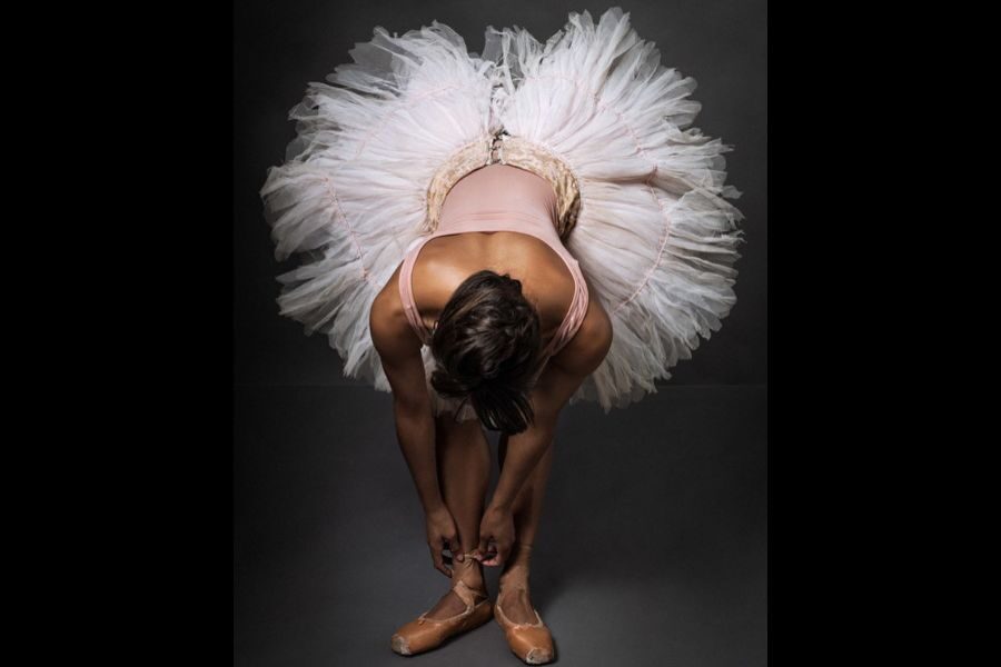 Ballet dancer tying ballet shoes, viewed from above. Wearing a fluffy white tutu and pink leotard, set against a dark background. Poised and graceful.