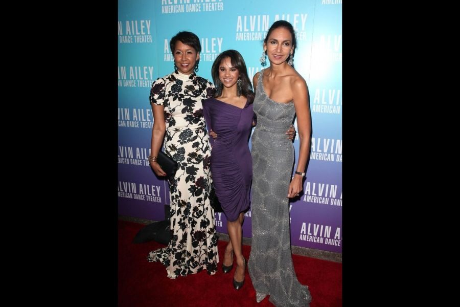 Three women pose on a red carpet at the Alvin Ailey American Dance Theater event. They wear elegant gowns, exuding confidence and joy.