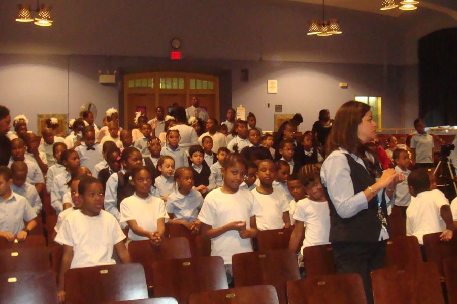 A group of young children in uniform stands in an auditorium, some seated, some standing, with a teacher guiding them. The mood is attentive and organized.