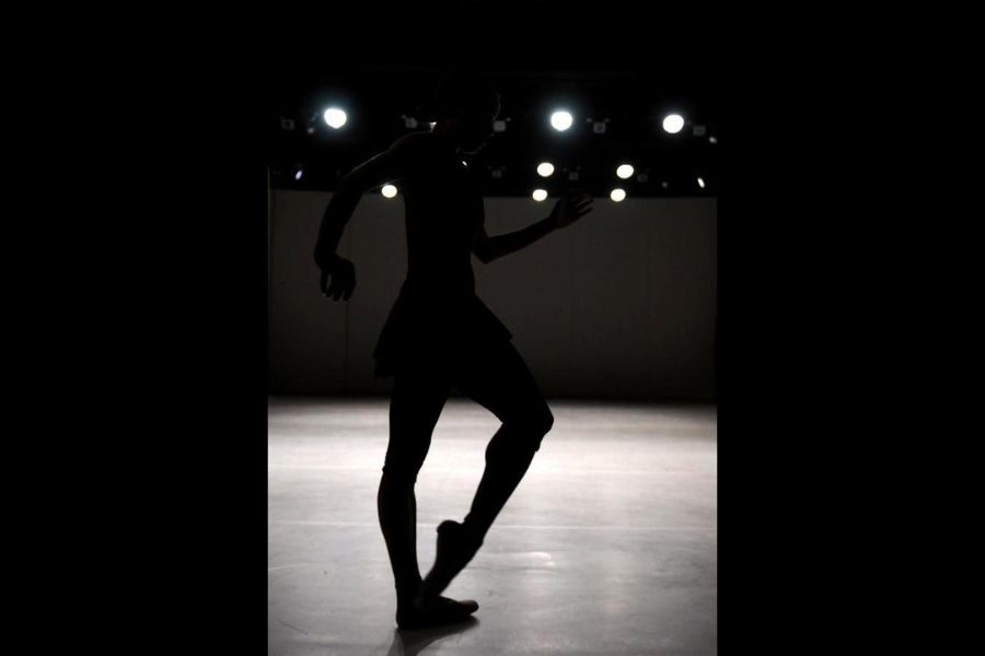 Silhouette of a dancer poised mid-motion in a dimly lit studio. Bright overhead lights create a dramatic contrast, highlighting graceful movement.
