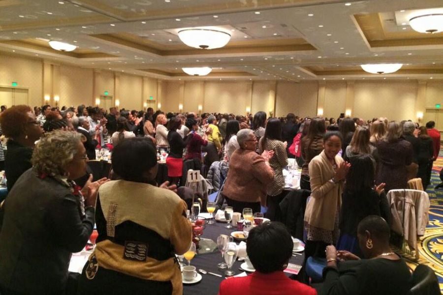 A large, diverse group of people stands and claps in a banquet hall. Tables are set with glasses and plates. The atmosphere is celebratory and energetic.