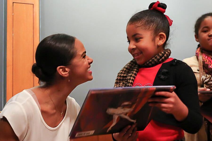 A smiling woman kneels to talk to a young girl holding a book with a ballet dancer on the cover. Another girl stands nearby, smiling warmly.