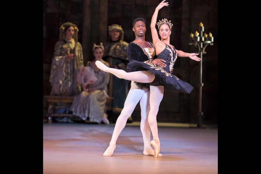 Ballet dancers in elegant costumes perform a poised duet on stage. The woman, in a black tutu and crown, stands en pointe, while the man supports her gracefully.