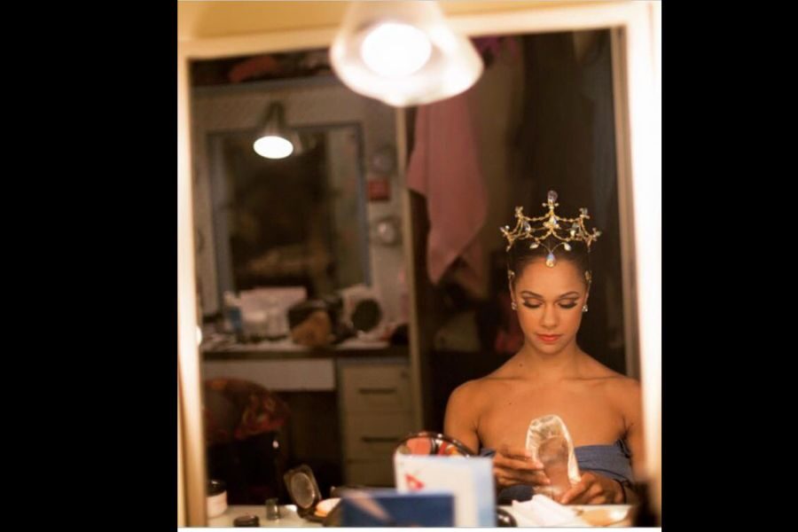 Ballet dancer preparing in a dressing room, wearing a decorative star-shaped headpiece. She holds a pointe shoe, looking focused under soft lighting.