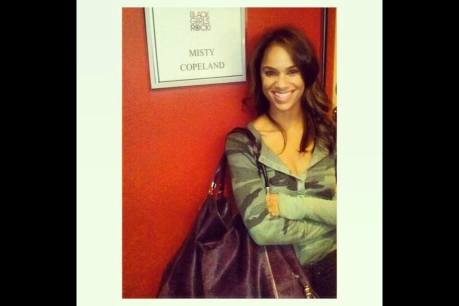 Smiling woman in a camouflage jacket stands in front of a red wall with a sign reading "Misty Copeland." She exudes confidence and warmth.
