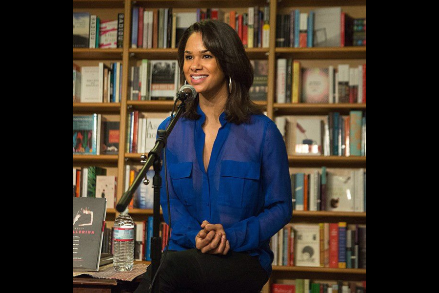A woman in a translucent blue blouse smiles while seated in front of a microphone. Bookshelves filled with books are in the background, creating a warm, intellectual ambiance.