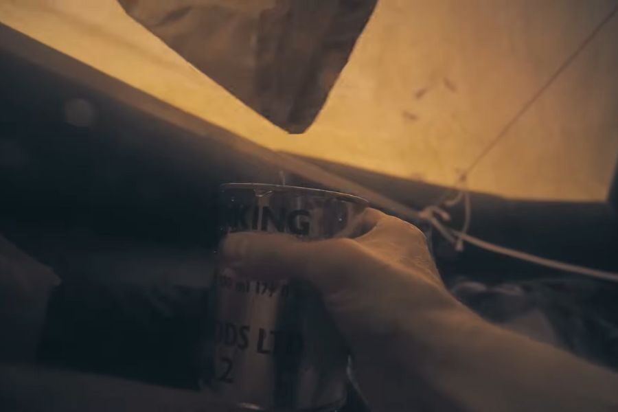 A hand pours liquid from a packet into a metal mug inside a softly lit tent. The scene conveys warmth and coziness in a camping setting.