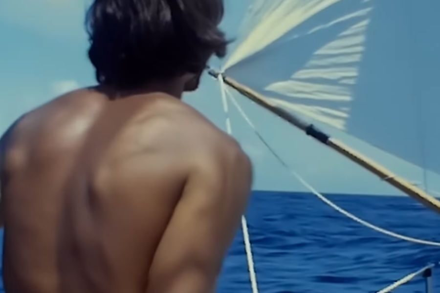A shirtless person with long hair is seen from behind, steering a sailboat in the open ocean. The sky is clear, and the sea is calm, providing a sense of adventure and freedom.
