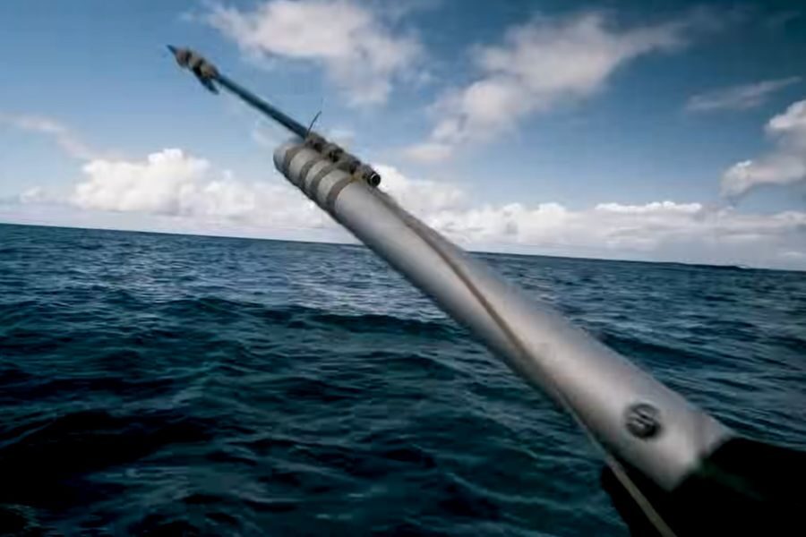 Close-up of a silver harpoon aiming over a vast, calm ocean beneath a blue sky with scattered clouds. The scene conveys adventure and focus.