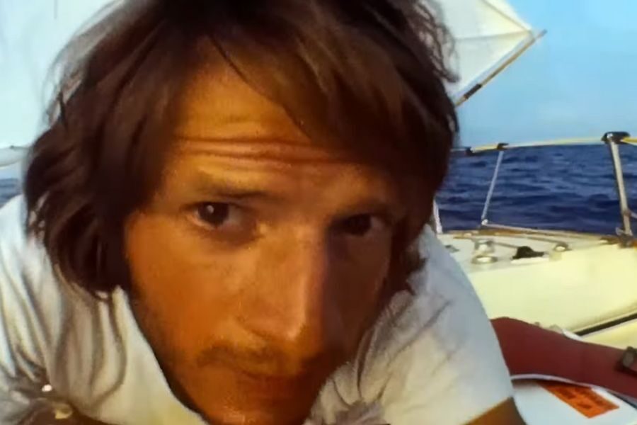 A man with tousled hair leans towards the camera on a sailboat, wearing a white shirt. The sea and sail are visible, suggesting a calm ocean setting.
