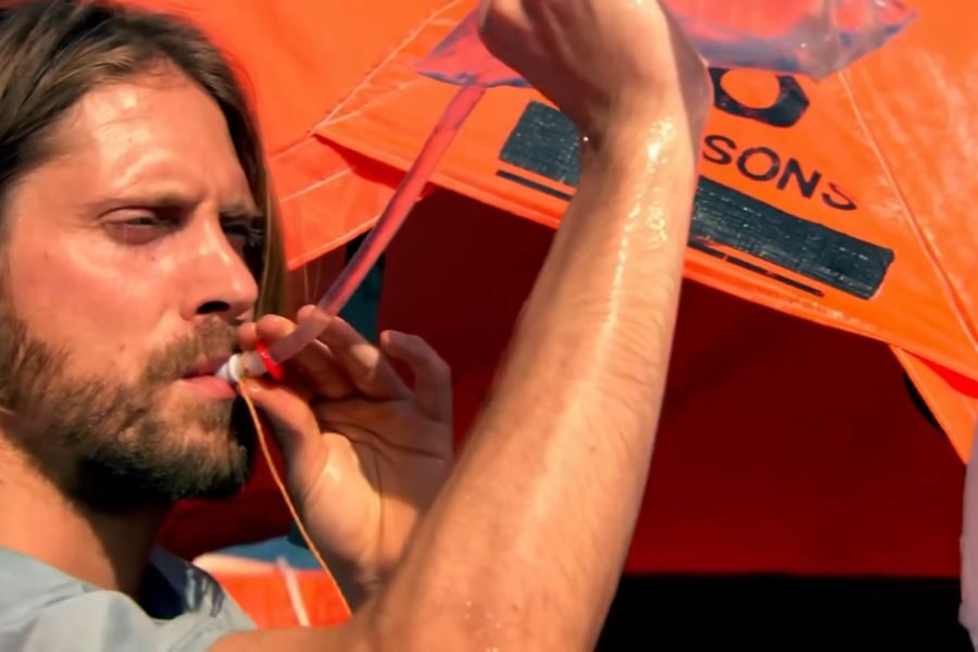 A man drinks water from a plastic bag with a straw, against the backdrop of a bright orange tent or lifeboat. He appears focused and determined.