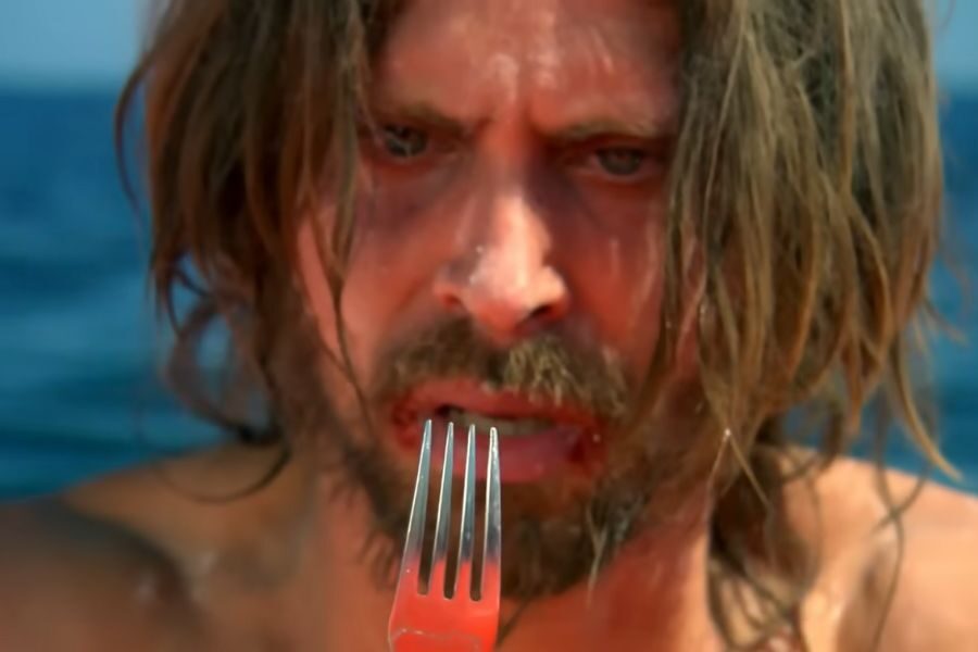 A distressed man with long hair and a beard holds a fork, staring intently. The backdrop is an expansive blue ocean, suggesting isolation.