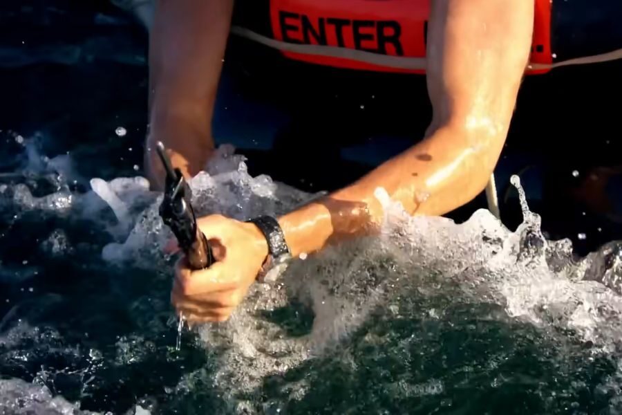 Close-up of a person's arms gripping a rope in turbulent water, wearing a red life vest. The scene conveys urgency and struggle, suggesting a survival context.