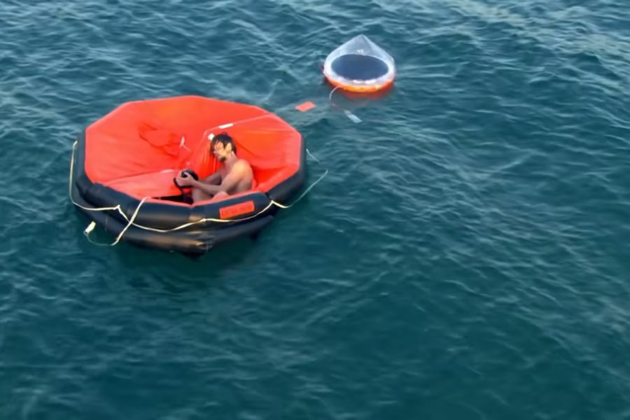 A man in a small, red inflatable raft floating on calm blue water, exhausted, pumping his deflating raft. A transparent circular buoy is tethered nearby.