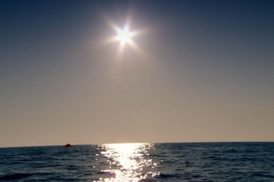 Bright sun in a clear sky reflecting on calm ocean waves, creating a serene and peaceful atmosphere. A small red raft is visible on the horizon.