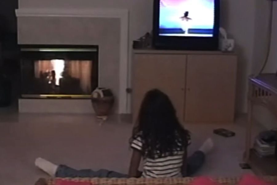 A child with curly hair, wearing a striped shirt and jeans, sits on the floor watching TV. A lit fireplace is to the left, creating a cozy atmosphere.