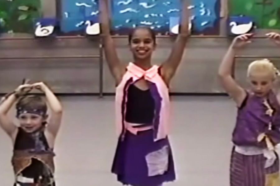 Three young children in colorful costumes pose with arms raised in a dance studio. They smile joyfully, and swan paintings are in the background.