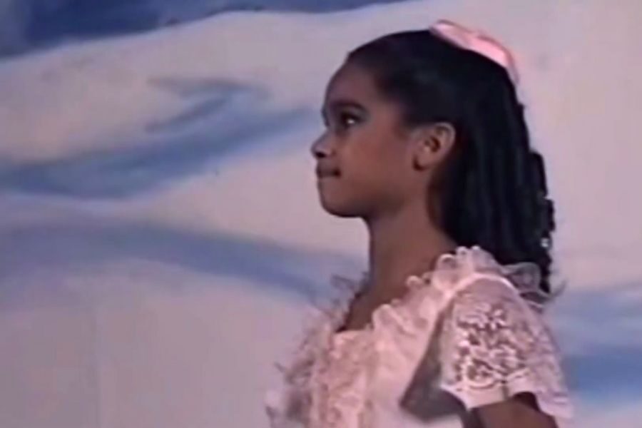 A young girl stands on stage in profile, wearing a lace dress and a pink bow. The background is softly blurred. She appears focused and serene.