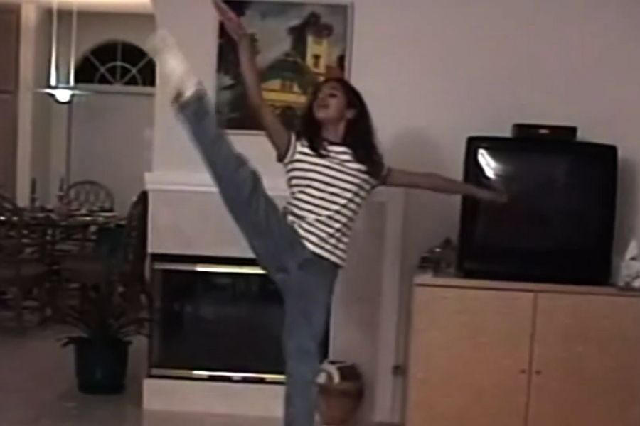 A person performs a high kick in a living room, wearing a striped shirt and jeans. The room has a TV, a painting, and a dining area. The scene feels lively.