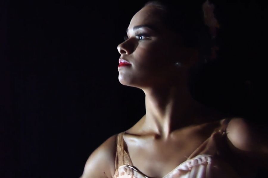 A dancer gazes confidently into the distance, her face partially lit, highlighting her red lipstick and elegant costume against a dark background.