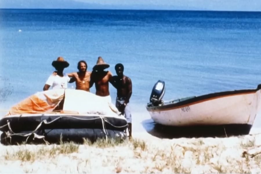 Four people stand on a sandy beach with two boats behind them, against a backdrop of calm blue sea and sky. They appear relaxed and content.