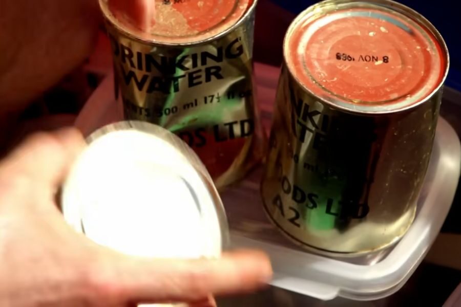 Two vintage cans labeled "Drinking Water" with red lids sit inside a plastic container. A hand is holding a metallic lid near the cans.