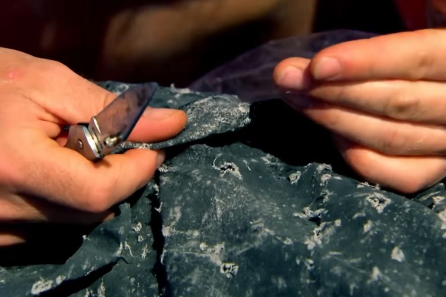 Close-up of hands using a knife to scrape a rough, textured surface covered in blue paint, creating a sense of focus and craftsmanship.