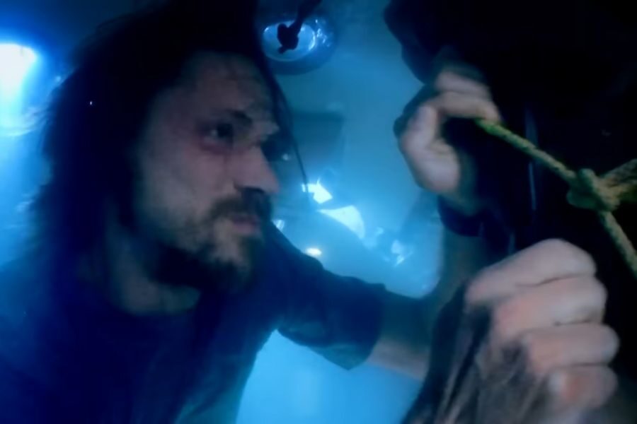 A man with wet hair is underwater, appearing tense and determined as he struggles with a knotted rope. Blue lighting creates an urgent, suspenseful atmosphere.