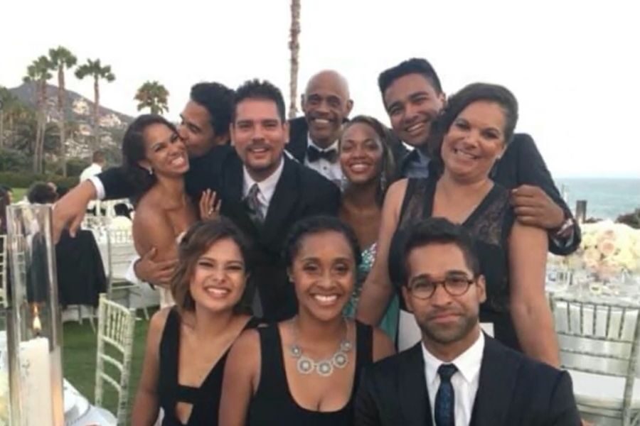 A group of ten people, dressed in formal attire, smiling at an outdoor event by the sea. Palm trees and tables are visible in the background.