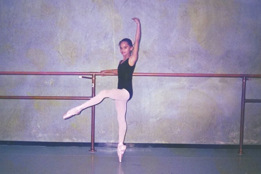 Young ballet dancer in black leotard and white tights practices at a barre, standing on one pointe shoe, arm elegantly raised, in a studio.