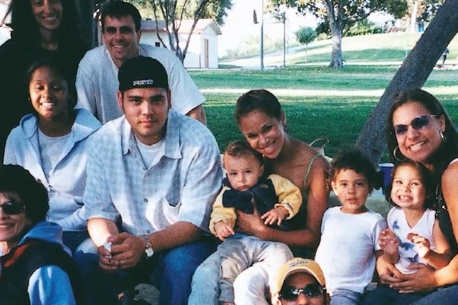 A group of adults and children sit closely together outdoors in a park, smiling warmly. Trees and a small building are in the sunny background, creating a cheerful atmosphere.