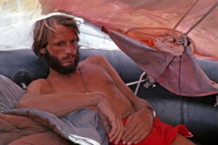 A shirtless man with a beard lies in a makeshift shelter on a raft, looking exhausted and pensive. There are pitched tarps and orange fabric overhead.