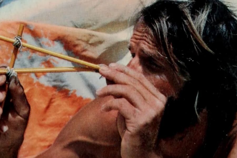 A person with long hair intensely focuses on holding a handmade slingshot, set against a colorful blurred background of orange and blue tones.