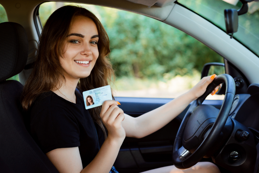 Smiling young woman sitting in the driver’s seat of a car, holding up her driver’s license with one hand on the steering wheel.